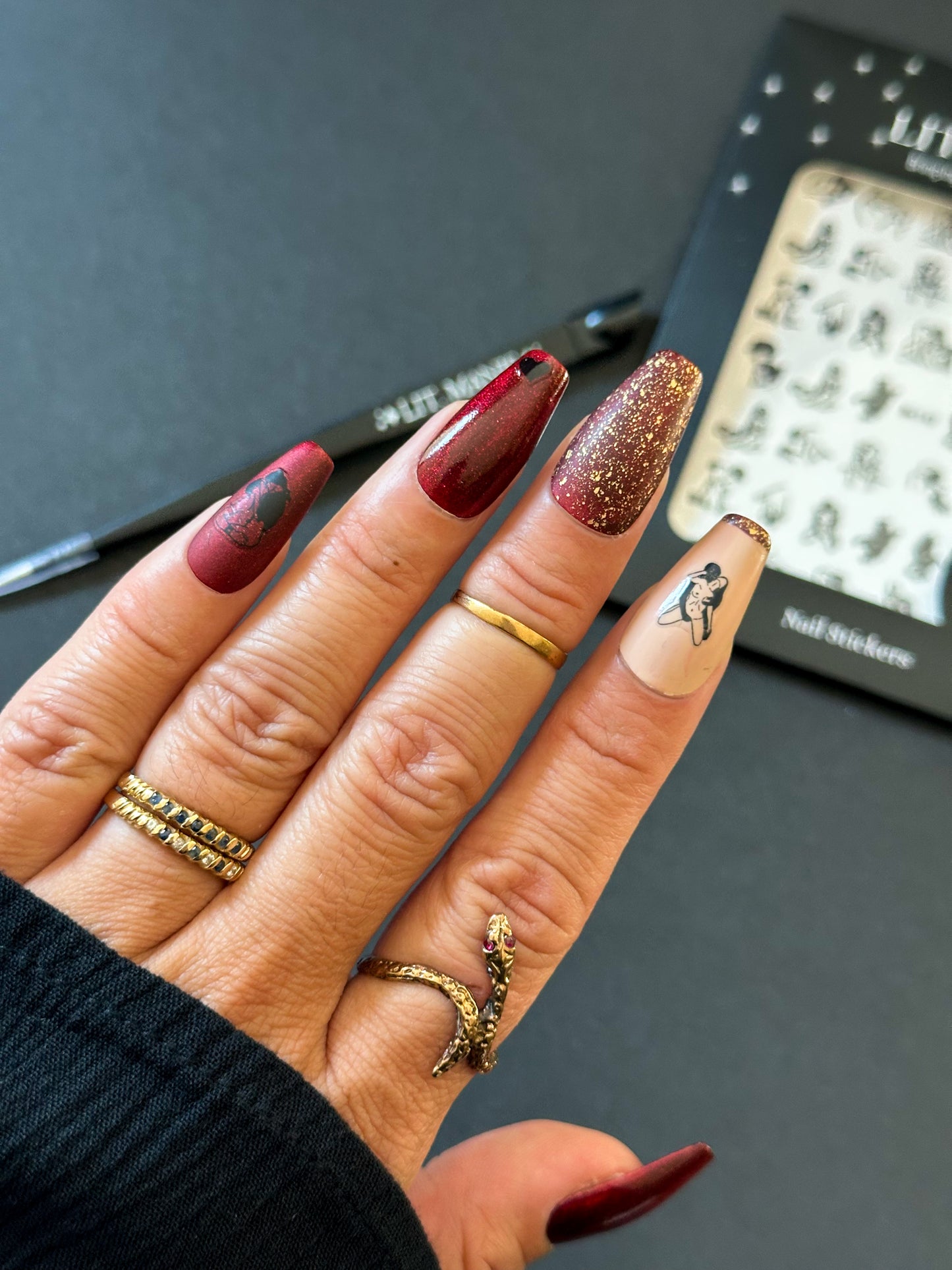 Hand with maroon and gold glitter nails, wearing gold rings, on a dark background.